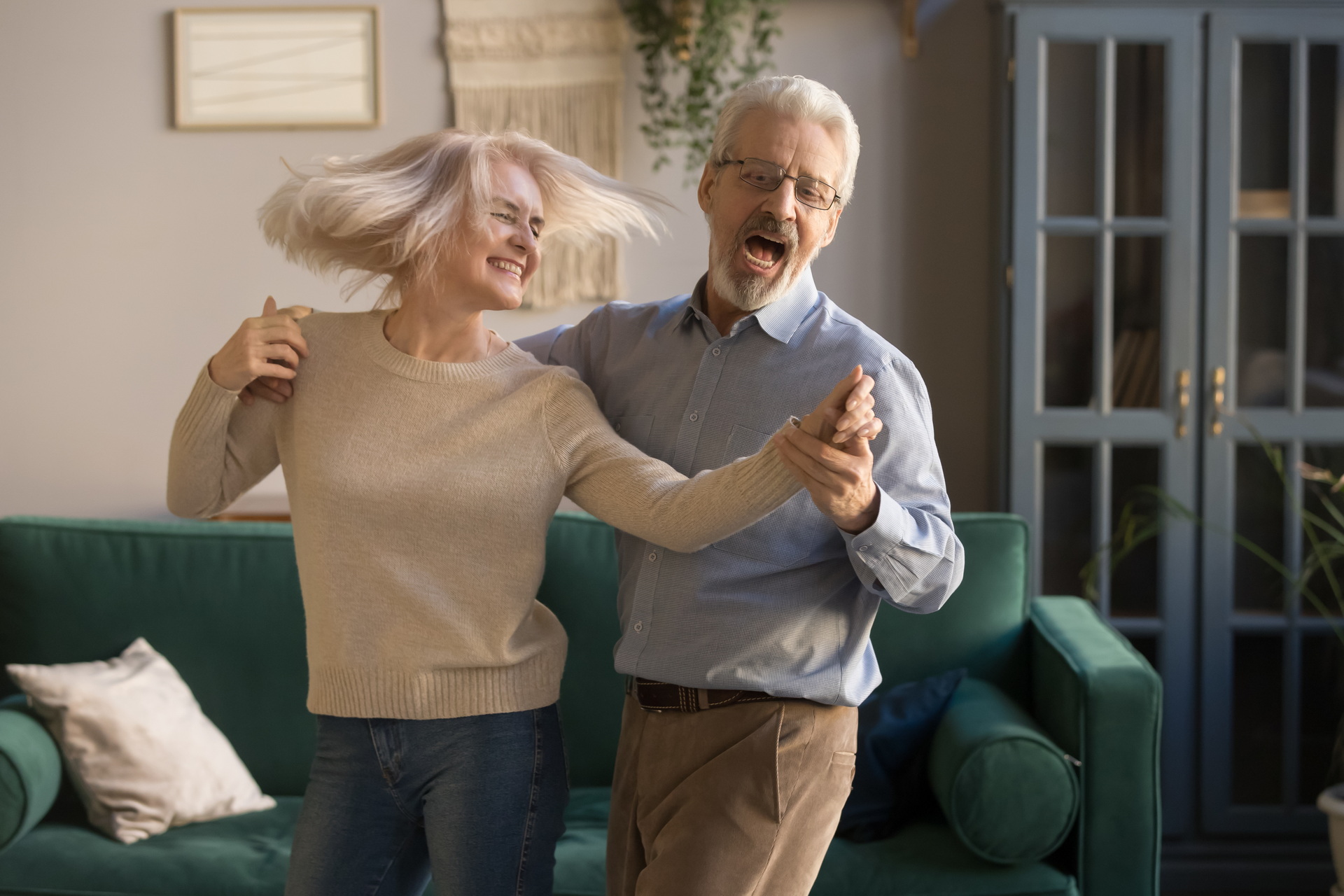 Carefree happy active old senior couple dancing jumping laughing in living room, cheerful retired elder husband holding hand of mature middle aged wife enjoy fun leisure retirement lifestyle at home