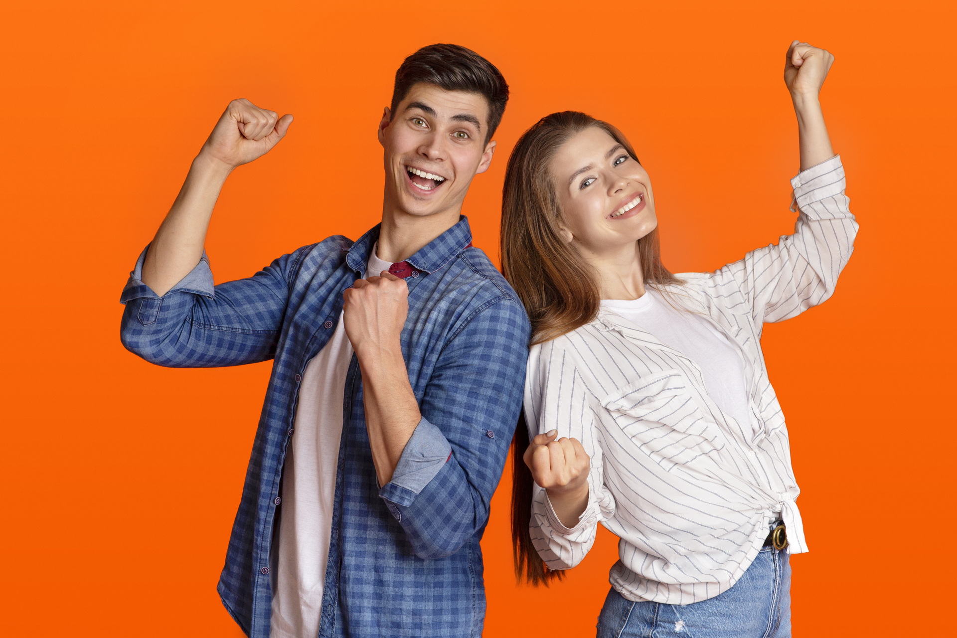 Success, dance and victory. Happy guy and lady in casual clothes celebrate winning, raise their fists up and express emotions of fun, isolated on orange background, studio shot, free space, cut out